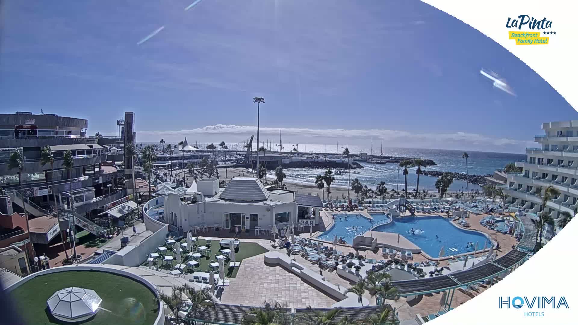A sunny day overlooking a resort with multiple pools and a beach, next to a marina and town.