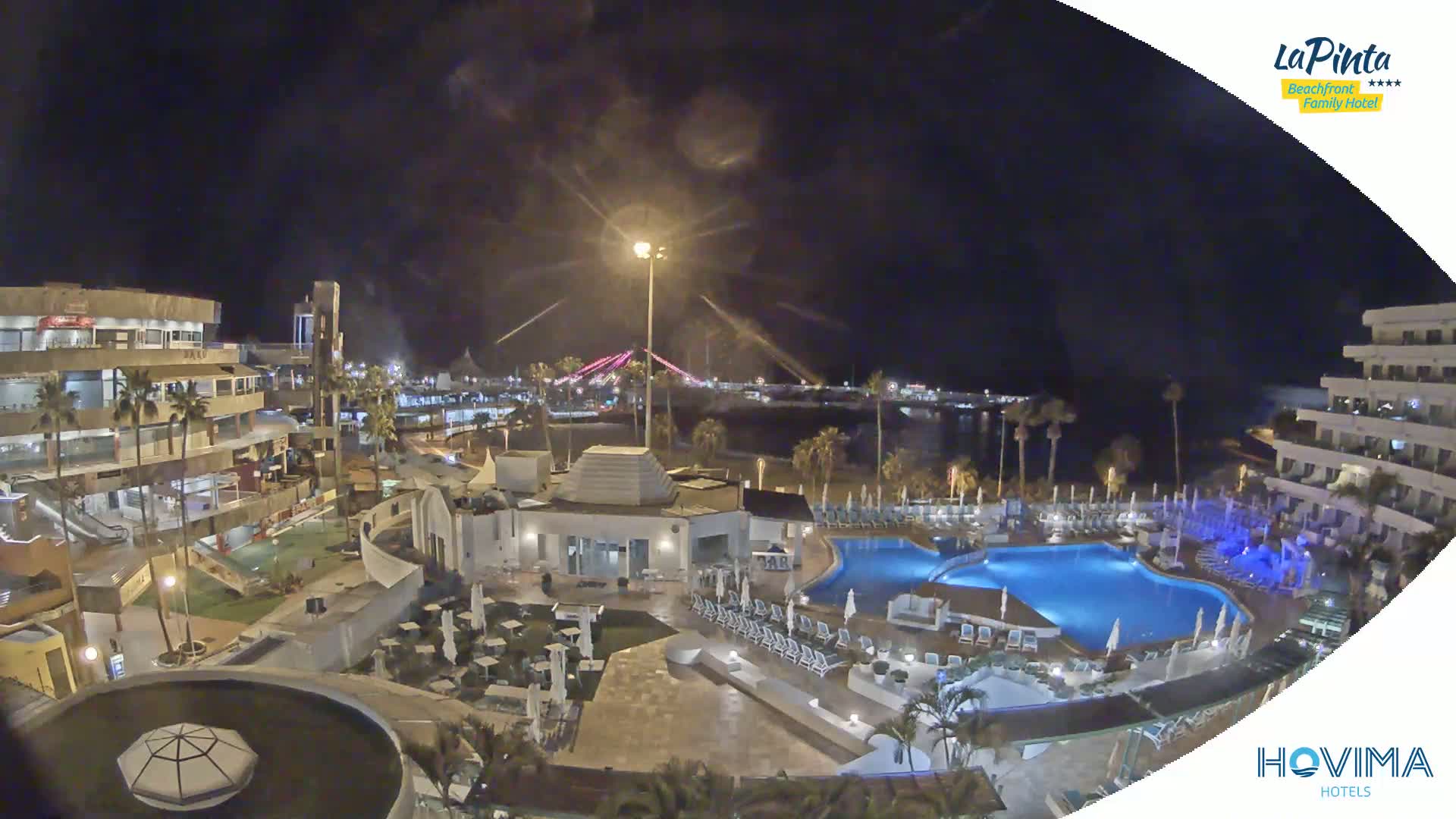 A nighttime aerial view of a resort complex with a swimming pool, situated near a beach and illuminated bridge under a clear, dark sky.