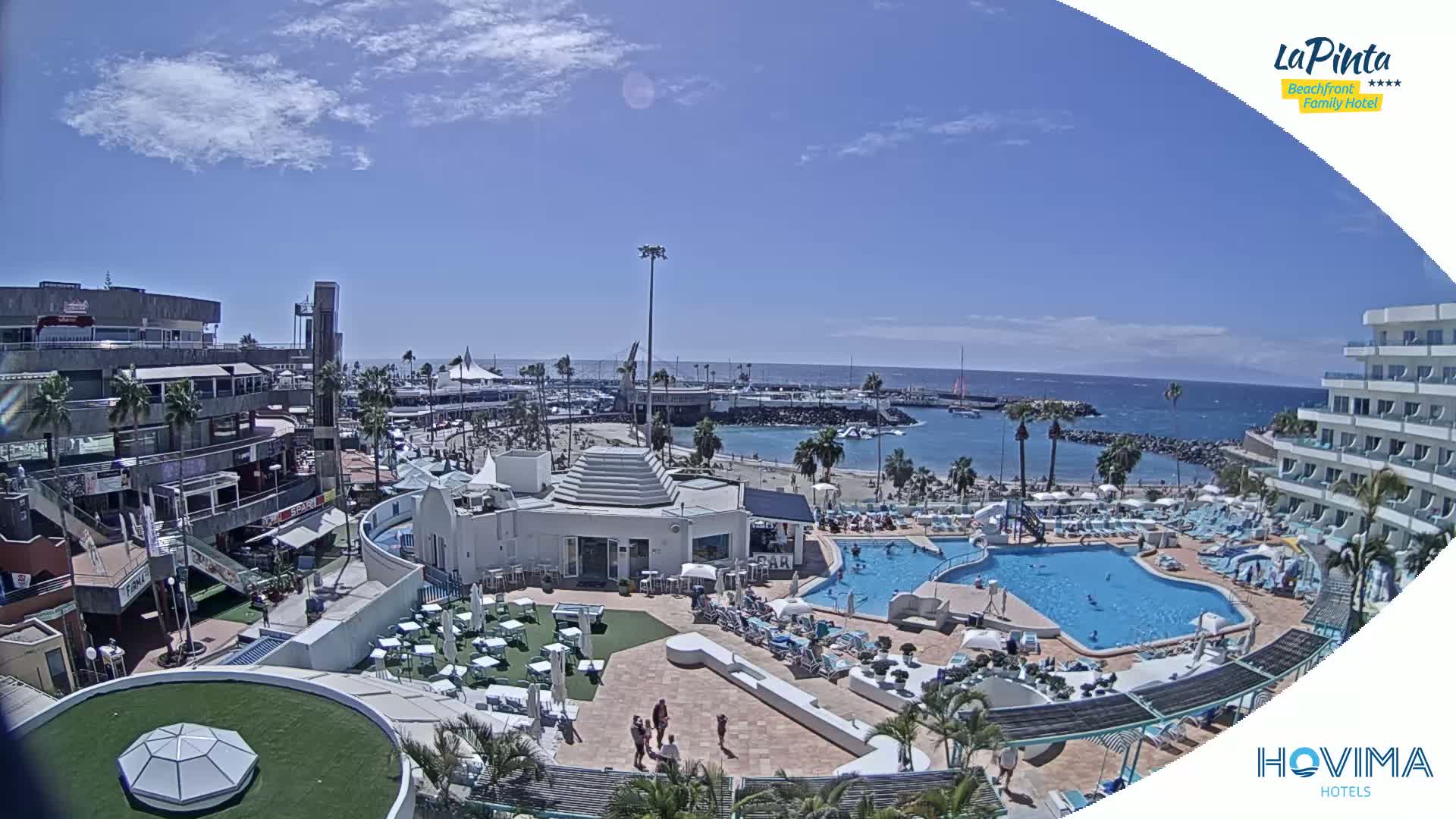 A sunny day at a resort overlooking a calm ocean, featuring a pool, beach, and buildings.