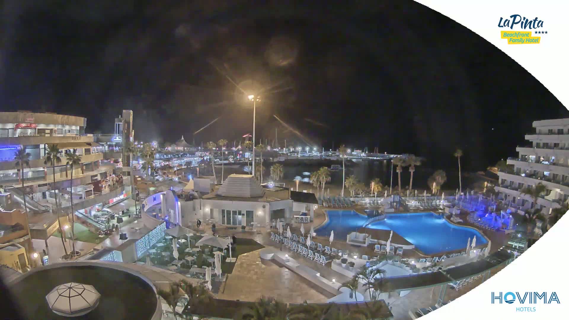 A nighttime aerial view of a resort complex with pools and surrounding buildings near a harbor under a clear, dark sky.
