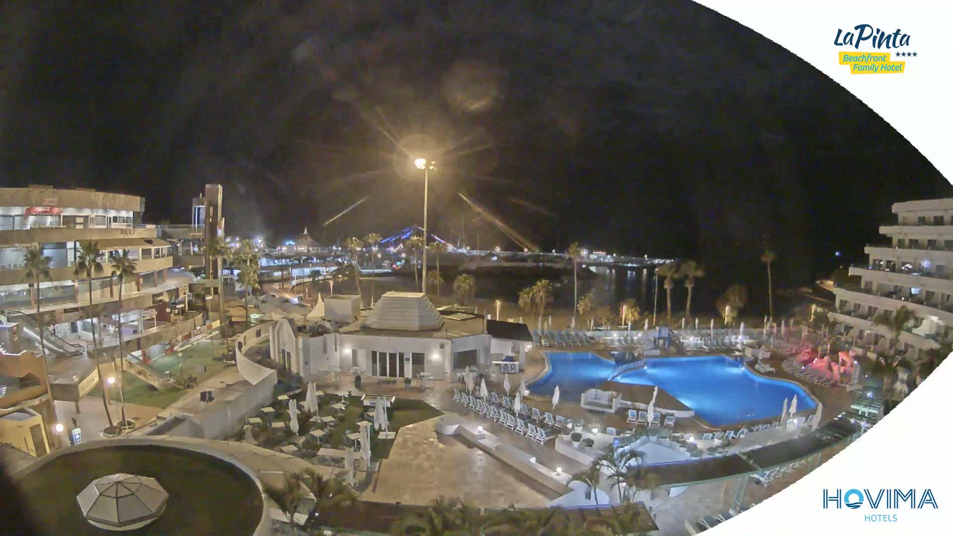 A nighttime aerial view of a resort complex with a swimming pool, situated near a waterfront area with palm trees and buildings under a dark sky.