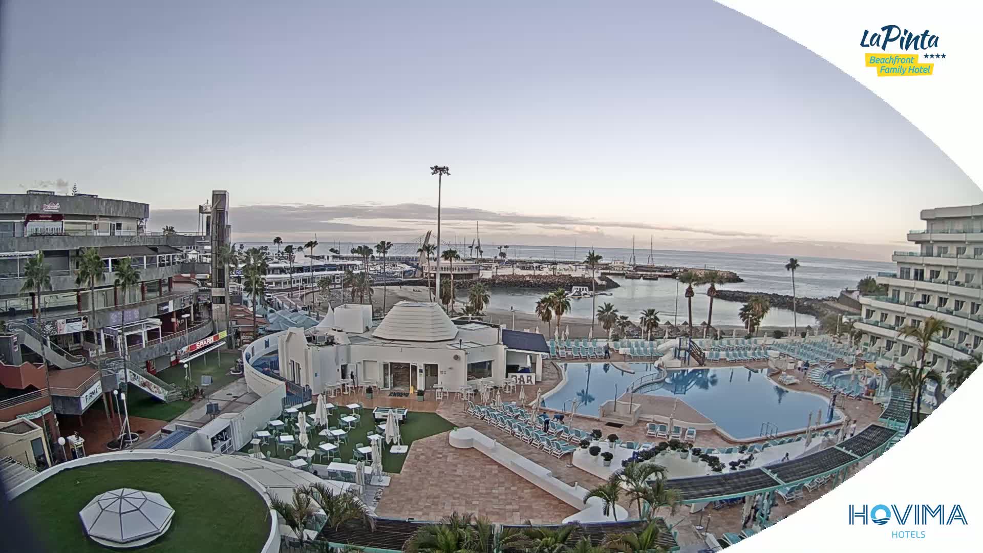 An elevated view shows a resort complex with swimming pools and a beachfront area overlooking a calm ocean under a clear, pale sky at dawn or dusk.