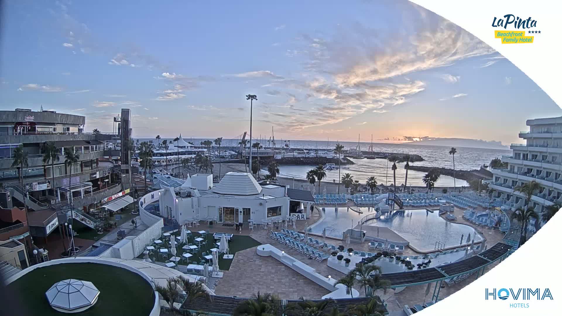 An aerial view of a resort at sunset overlooking a calm ocean and harbor, with a swimming pool and surrounding buildings in the foreground.