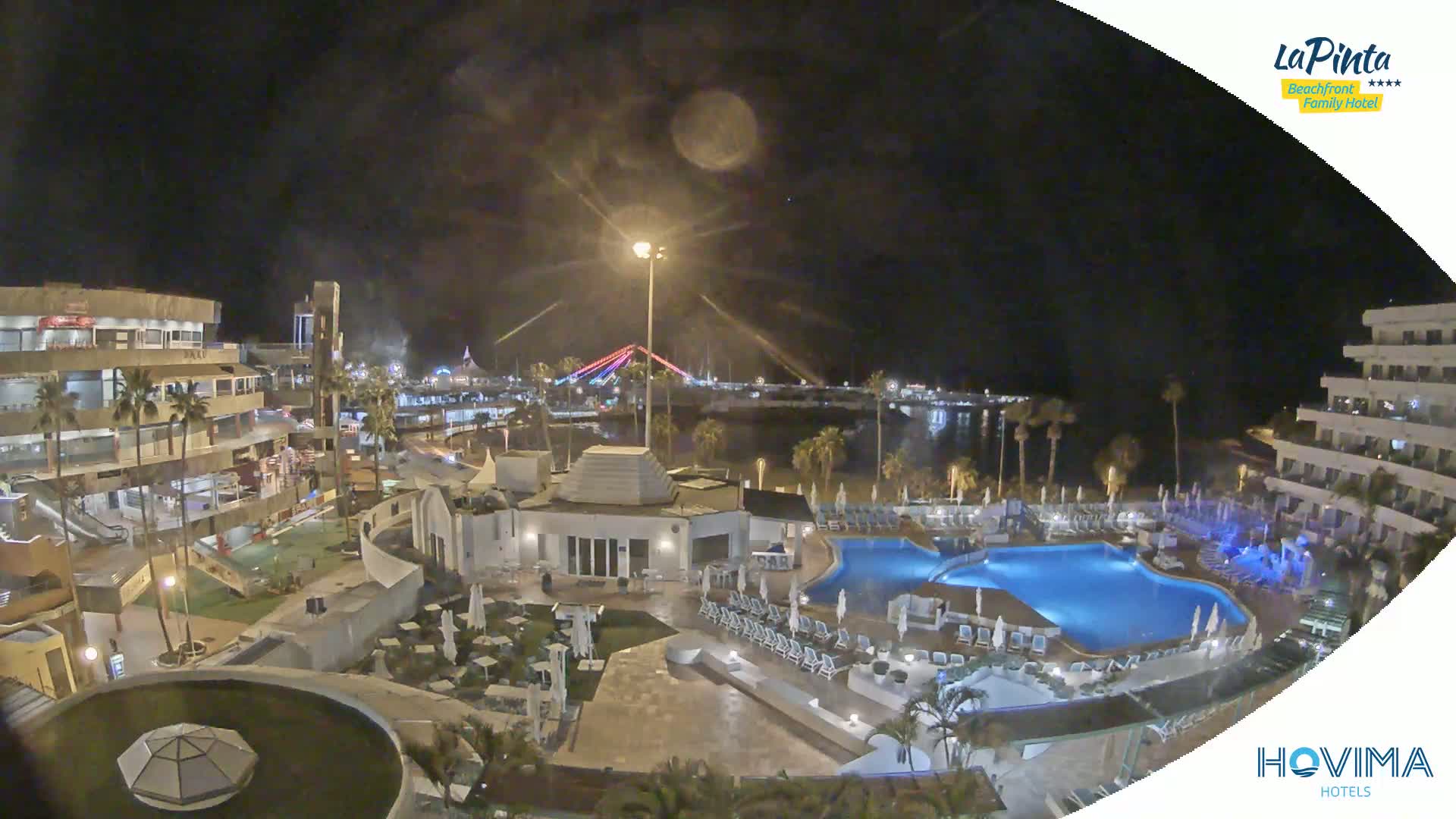 A nighttime aerial view of a resort complex with pools and surrounding buildings, overlooking a calm body of water and a bridge illuminated with lights.
