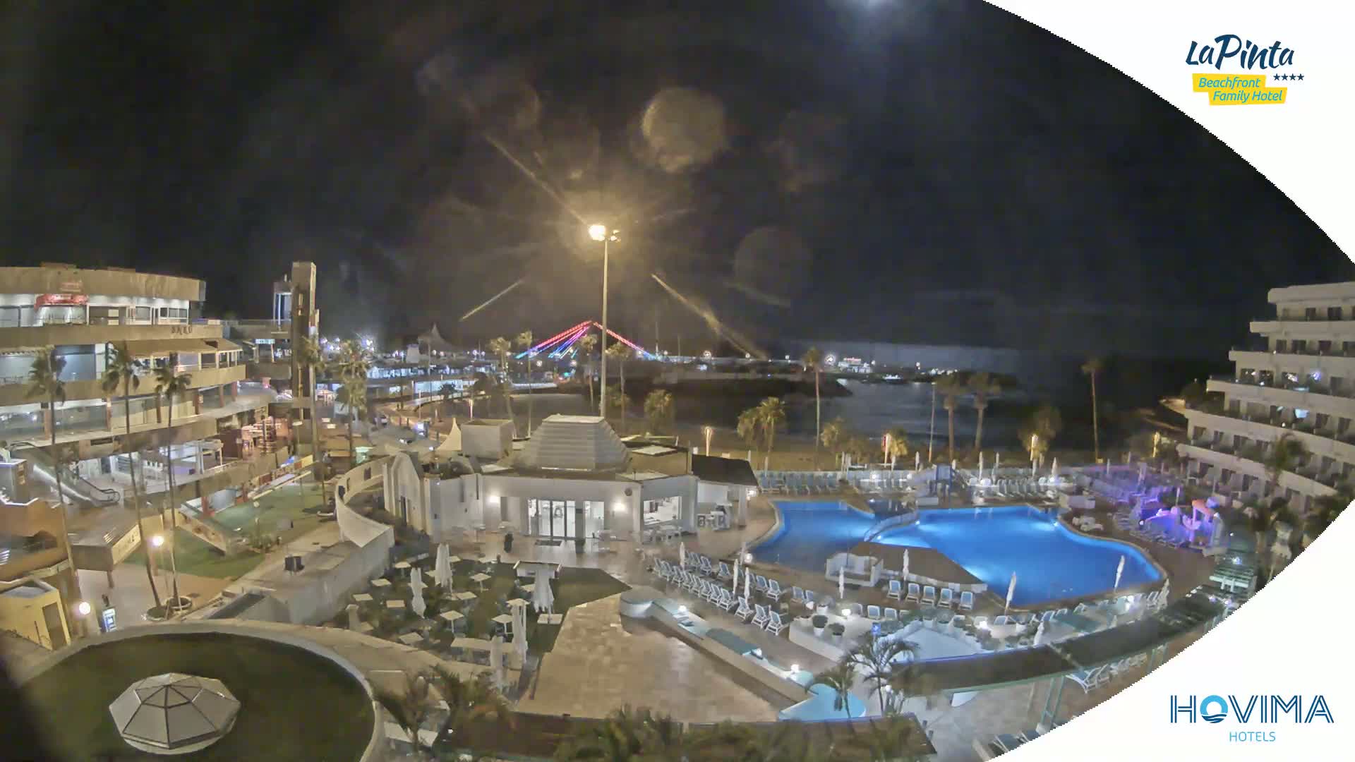 A nighttime aerial view of a resort complex with multiple pools, surrounding buildings, and a bridge in the distance under a partly cloudy sky.