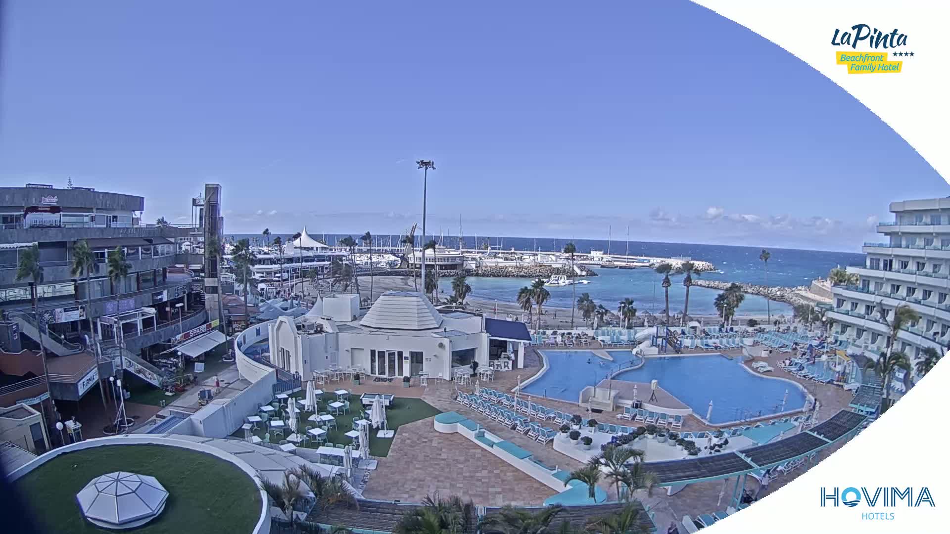 An aerial view shows a resort with a pool and surrounding buildings overlooking a calm ocean on a clear day.
