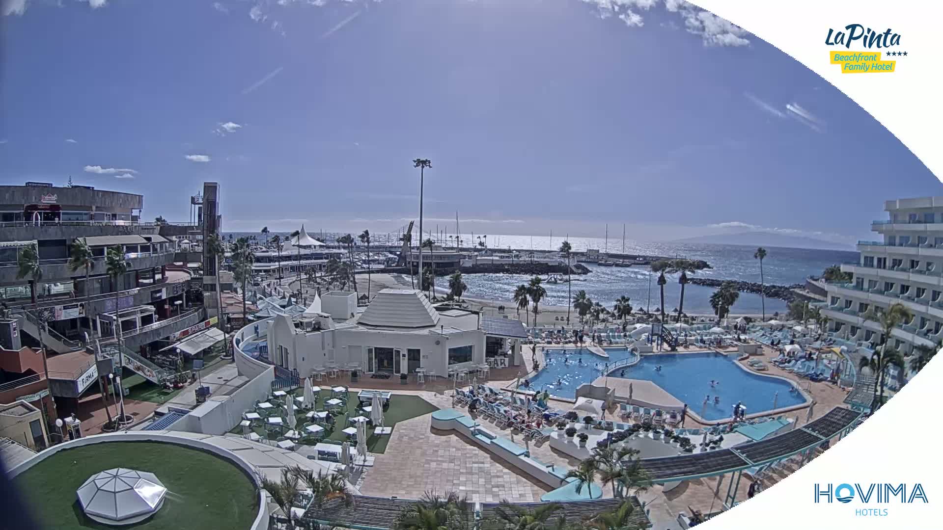 A sunny day shows a resort with a swimming pool and beach area overlooking a calm ocean with boats in a harbor.
