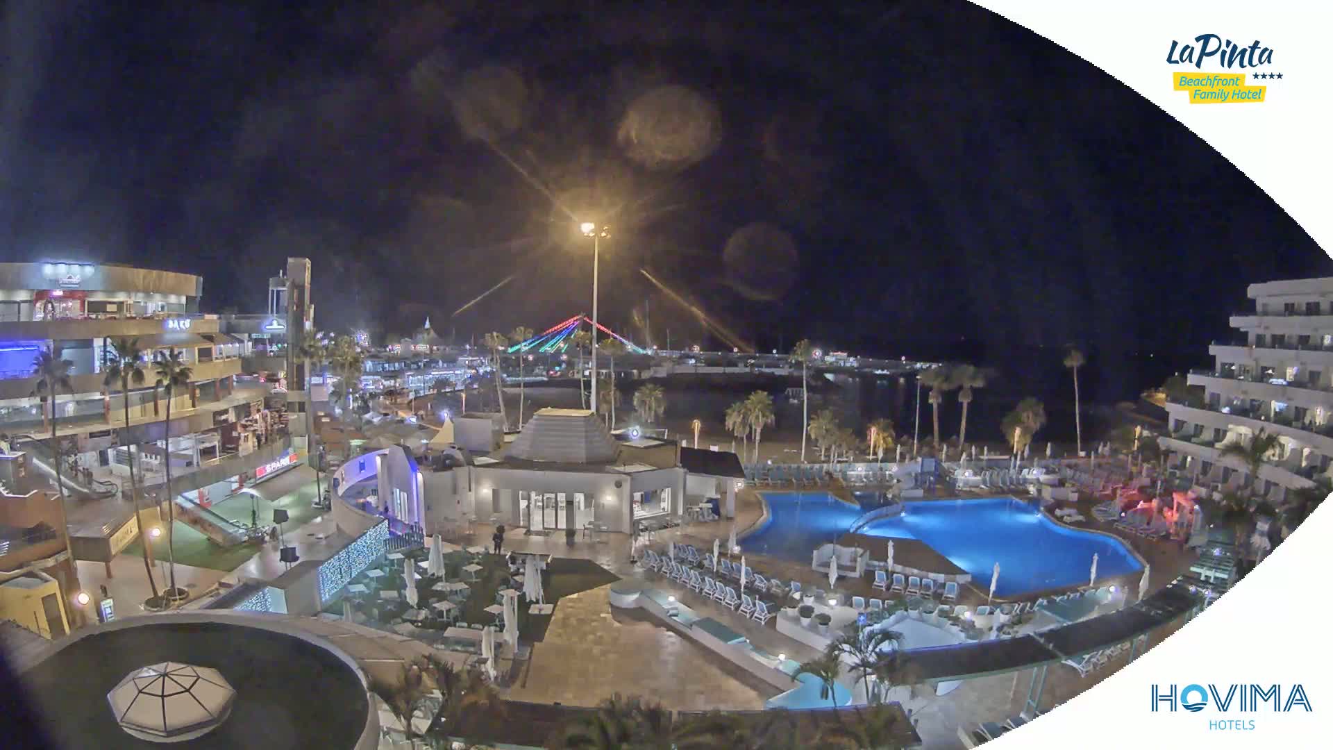 A nighttime aerial view of a resort with a pool and surrounding buildings, situated near a beach and a bridge with decorative lights.