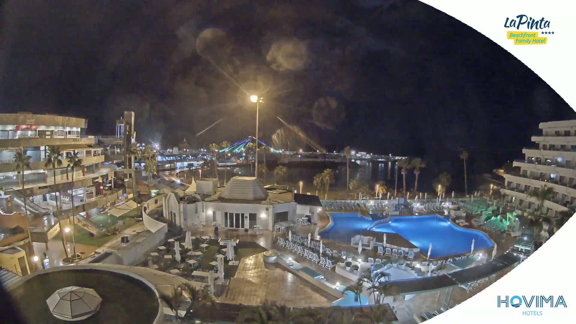 A nighttime view of a resort complex with a swimming pool, illuminated buildings, and a bridge in the distance under a partly cloudy sky.
