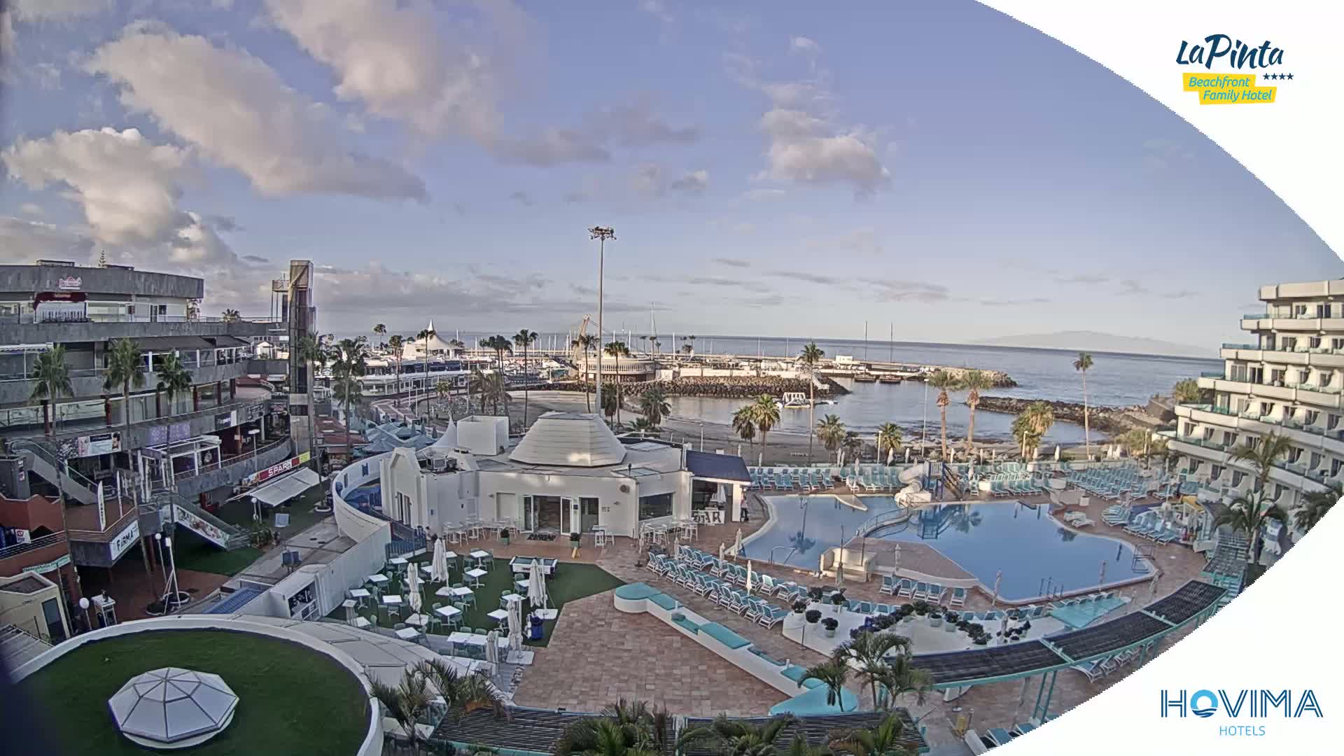 A resort with a swimming pool overlooks a harbor and calm ocean under a partly cloudy sky.