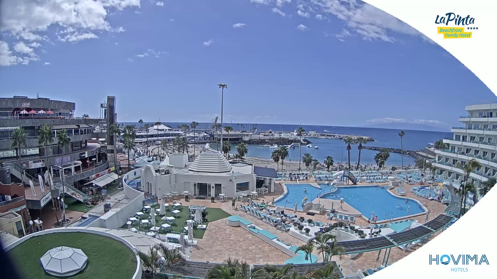 A sunny day overlooking a resort with a swimming pool complex next to a beach and harbor.