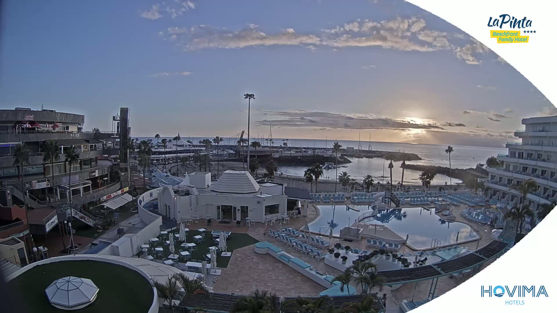 An aerial view of a resort at sunset overlooking a calm ocean, marina, and shops, under a partly cloudy sky.