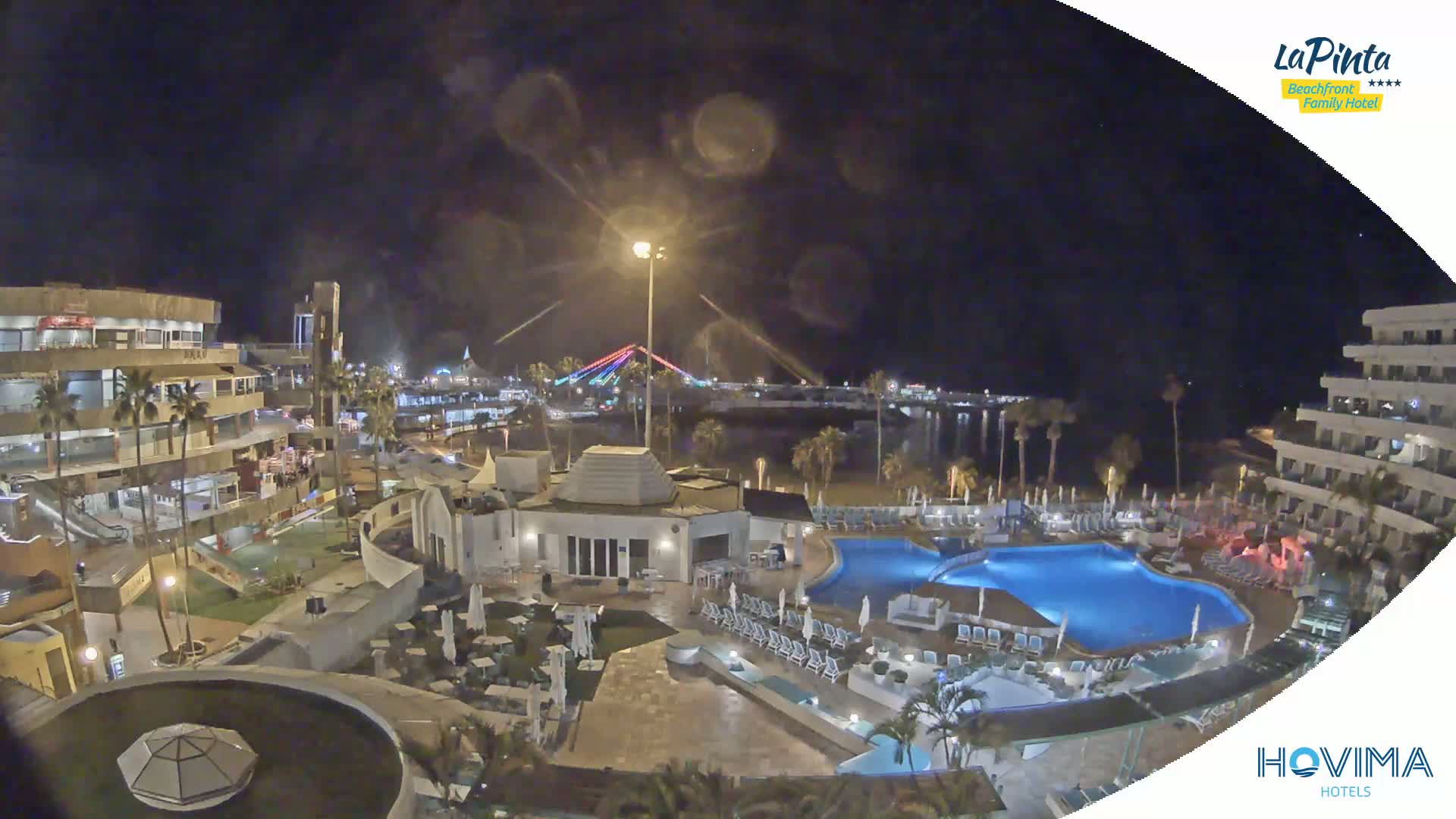 A nighttime aerial view of a resort complex with swimming pools, surrounding buildings, and a brightly lit bridge in the distance.