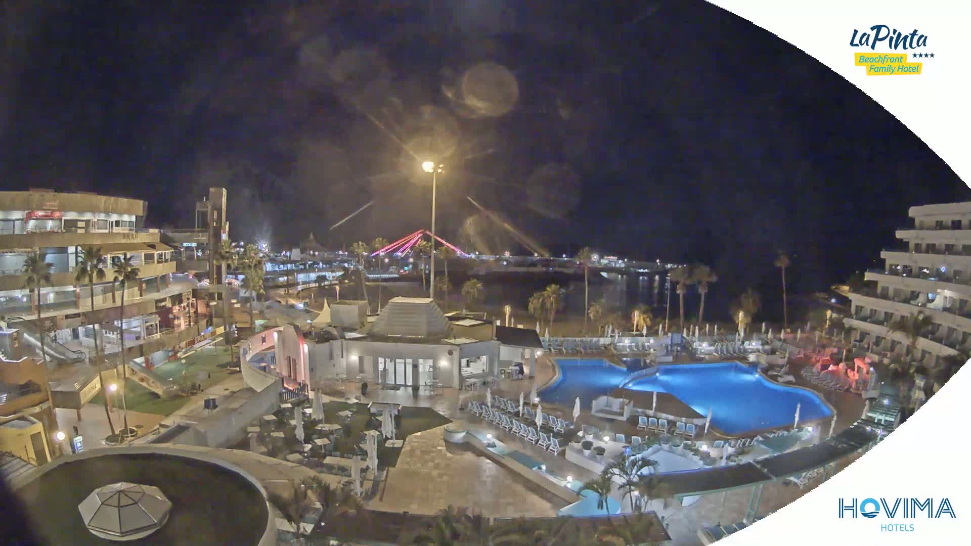An illuminated resort complex with multiple pools at night, overlooking a beach and a bridge lit with colored lights.