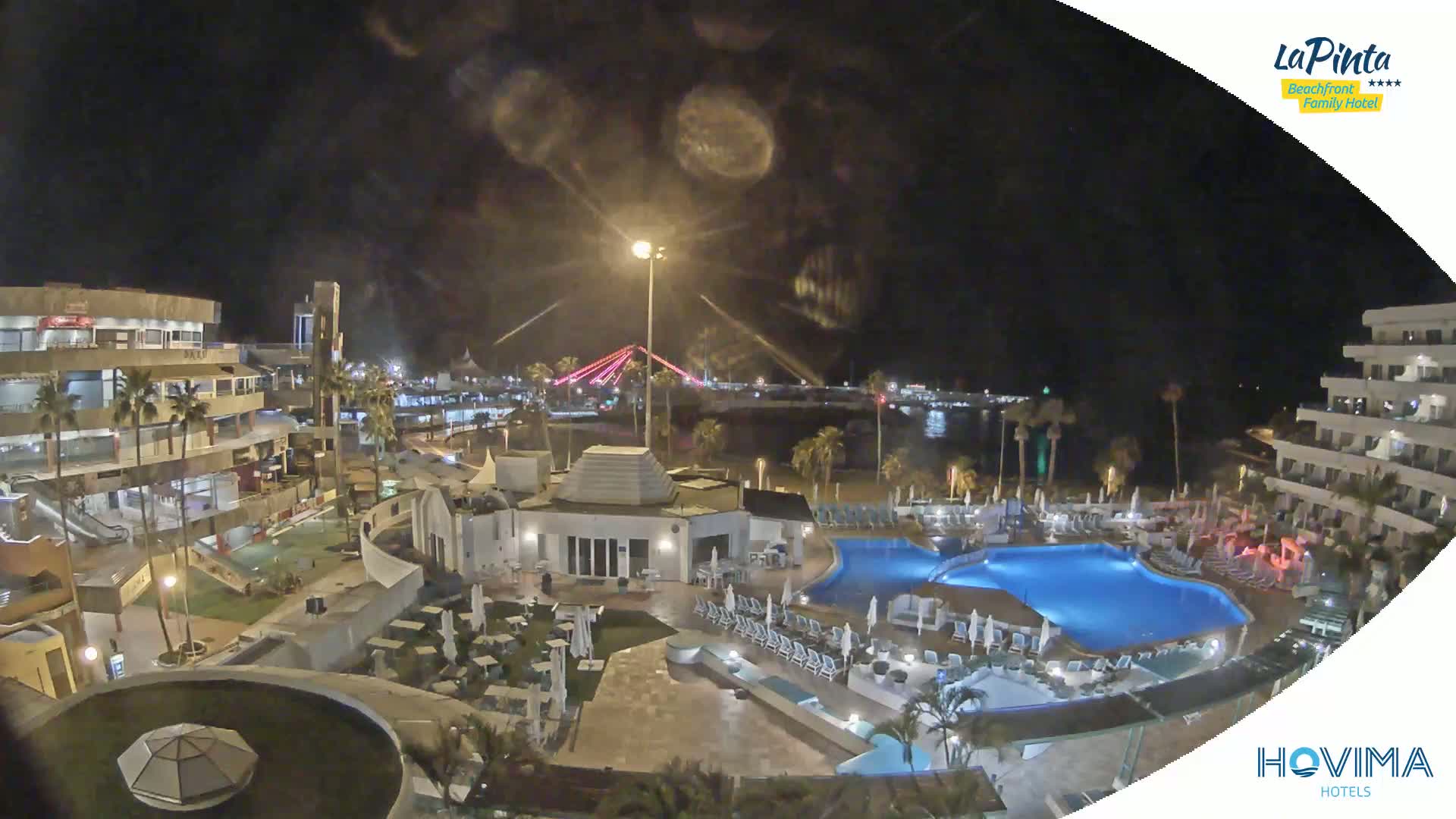 A nighttime aerial view of a resort complex with swimming pools, surrounding buildings, and a bridge illuminated in the distance.