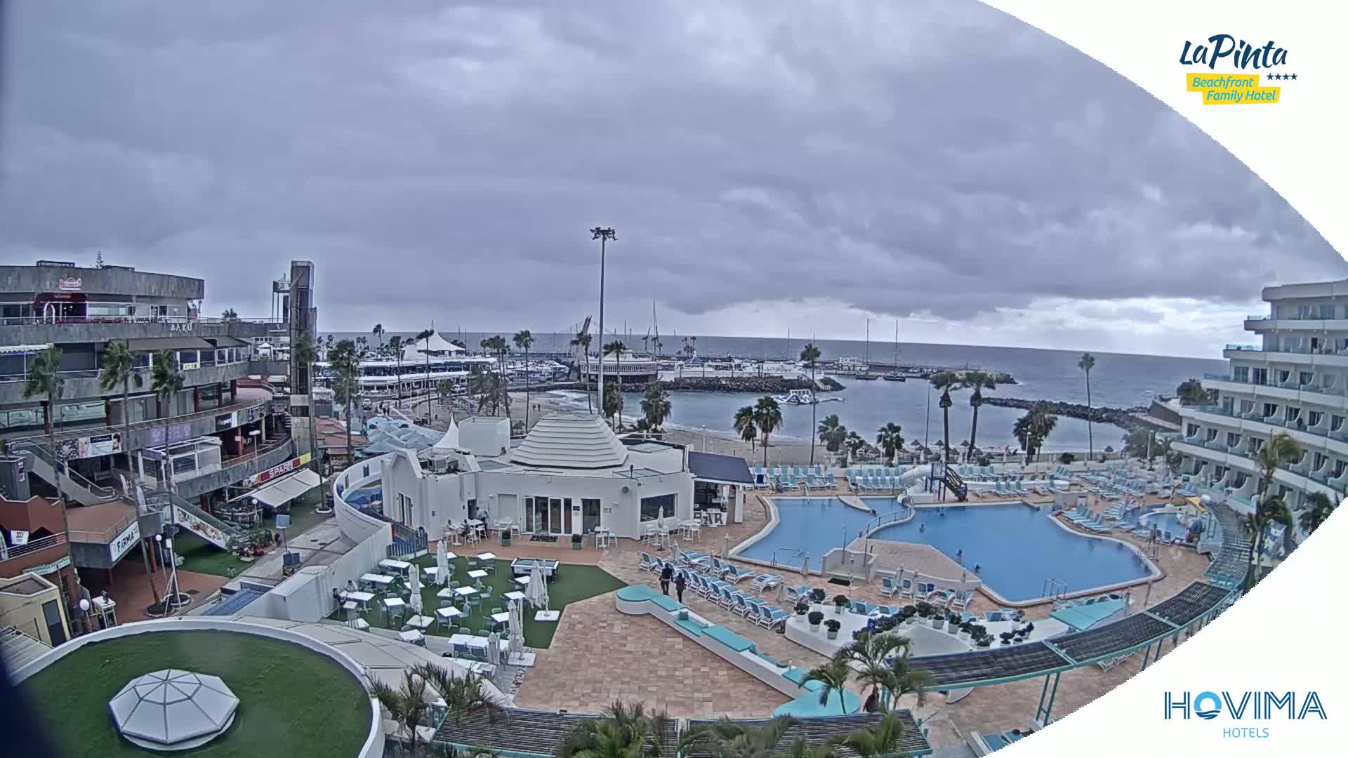 An overcast day shows a resort with a swimming pool and beach area overlooking a harbor, with shops and buildings nearby.