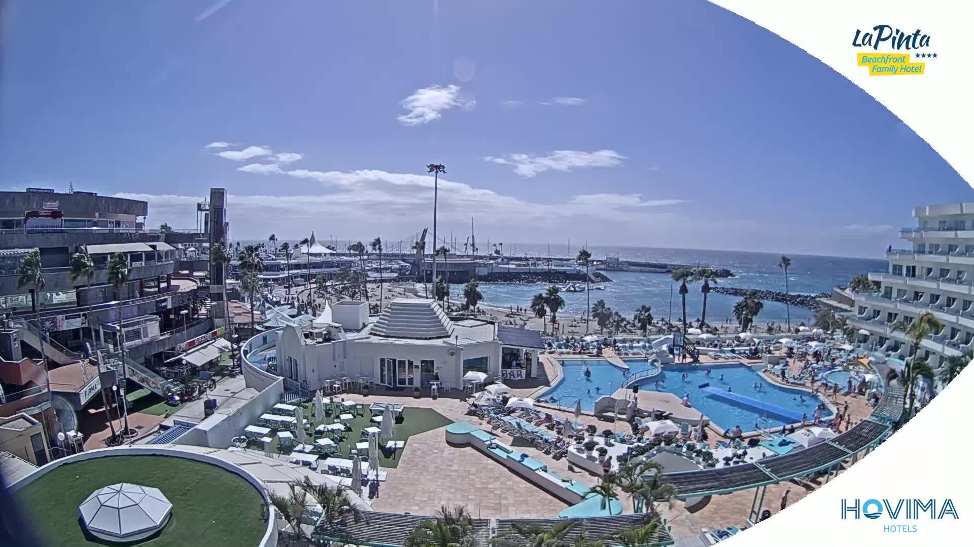 A sunny day at a resort shows multiple pools, a beach, and a marina with boats, all overlooking a calm ocean.