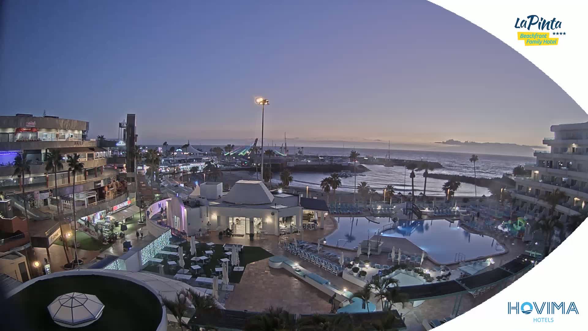 An aerial evening view shows a resort area with a pool, buildings, and a beach near a calm ocean under a twilight sky.