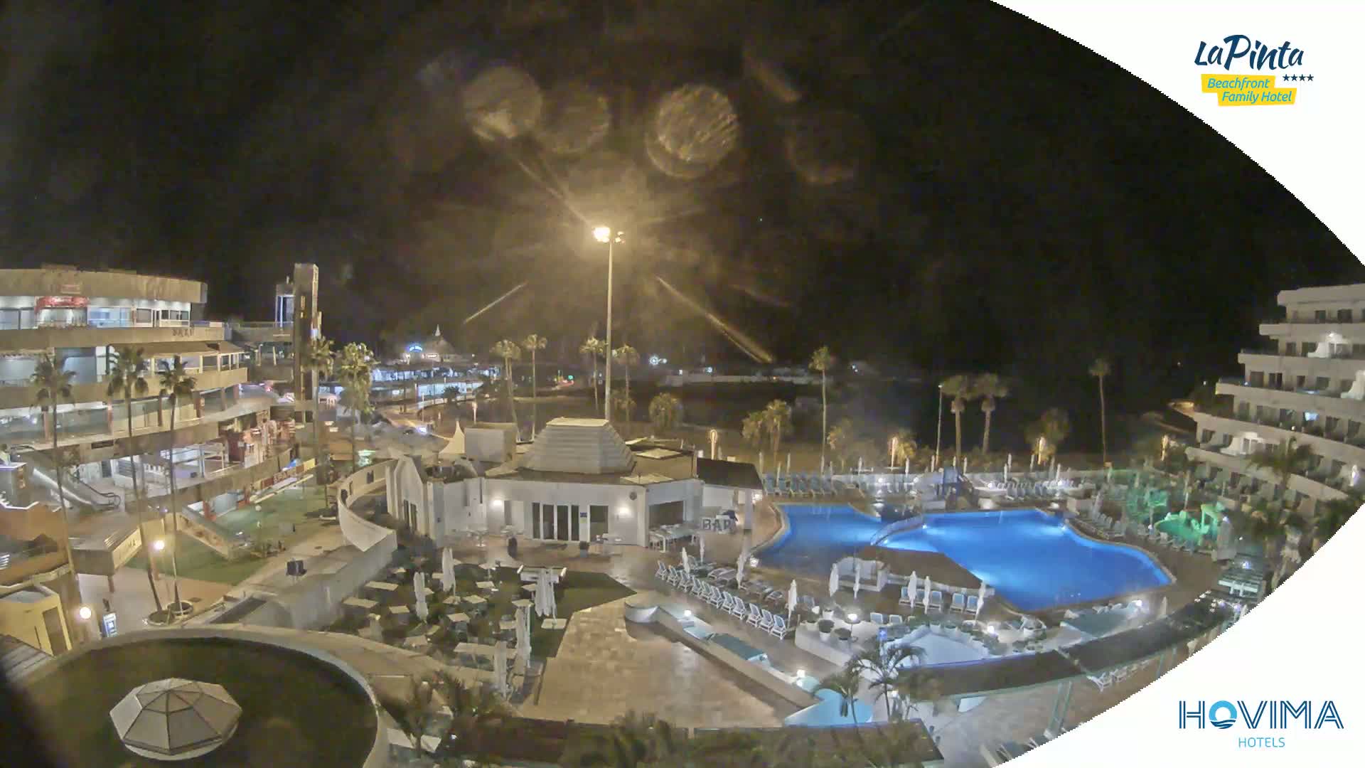 A nighttime, high-angle view of a resort complex with swimming pools, outdoor seating areas, and buildings, illuminated under a dark sky.
