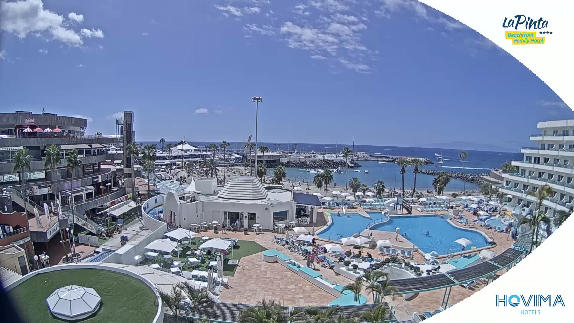 An aerial view of a resort area on a sunny day, showing a beach, pools, and buildings, with calm ocean waters in the background.