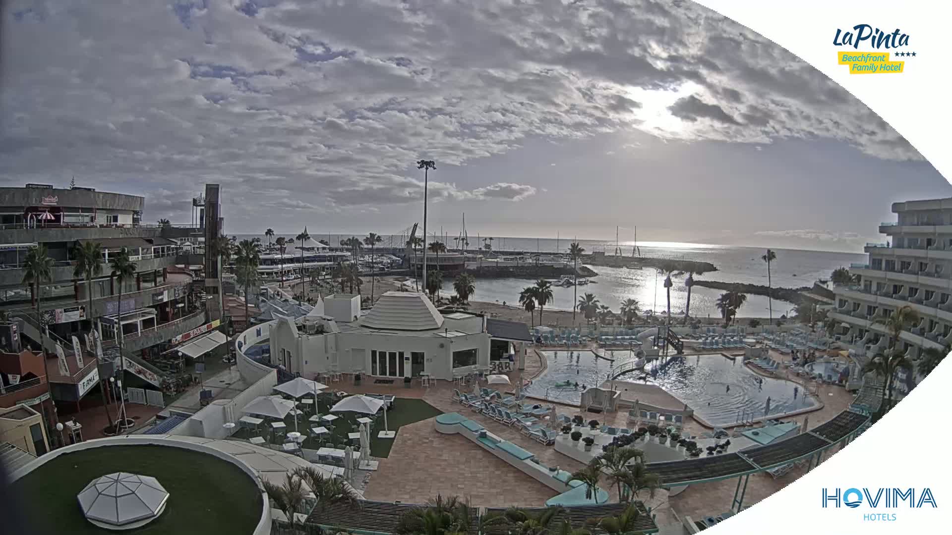 A resort area with a pool and beach, near a marina, under a partly cloudy sky.