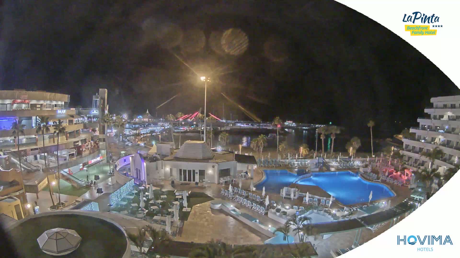 A nighttime view of a resort area with palm trees, swimming pools, and buildings illuminated near a body of water and a bridge.