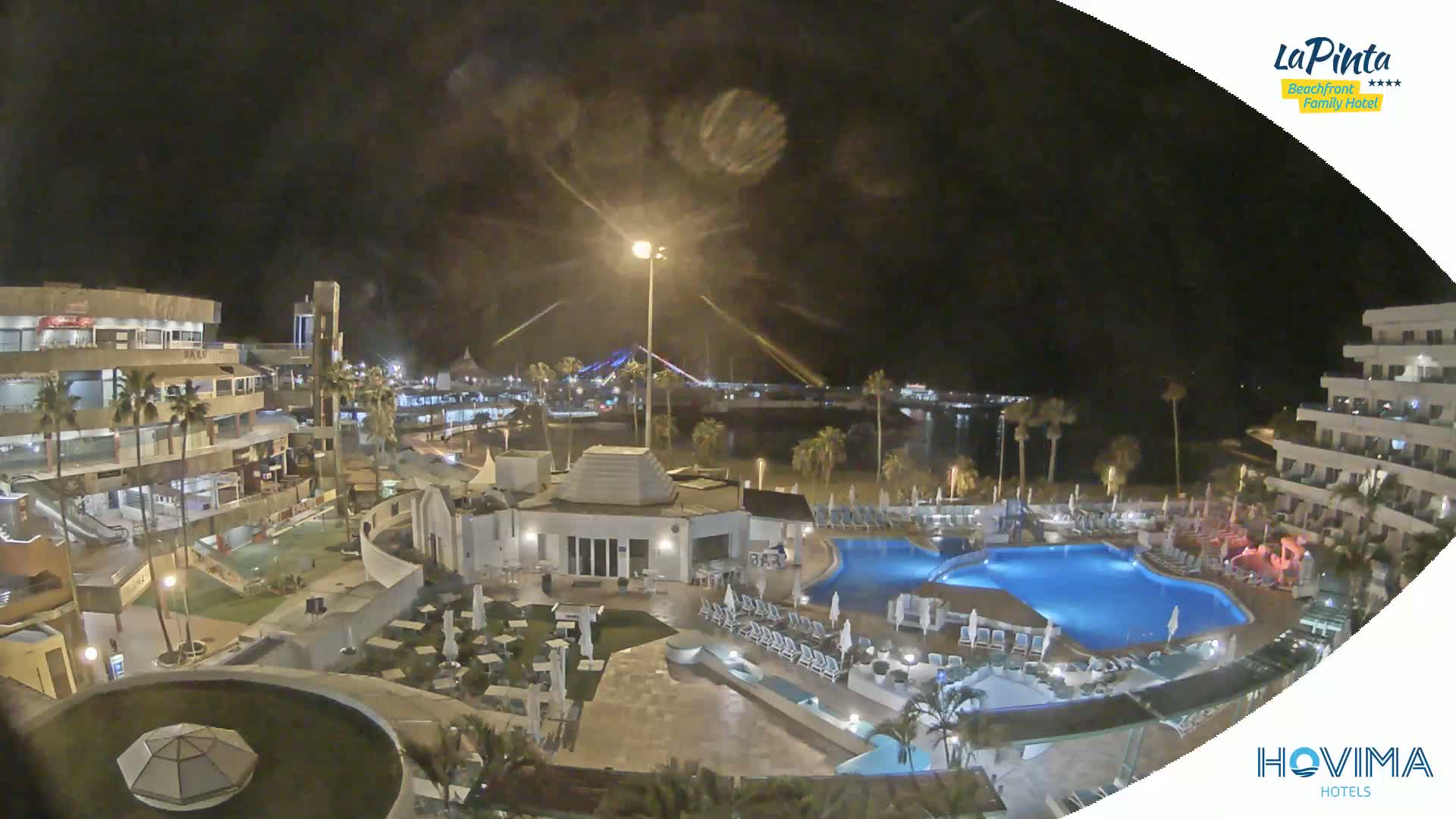 A nighttime aerial view of a resort complex with a swimming pool, surrounding buildings, and palm trees, overlooking a calm body of water.