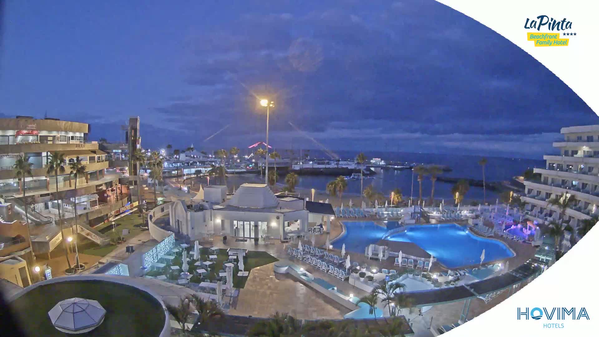 A nighttime view of a resort with a swimming pool complex, situated near a harbor and beach under a twilight sky.