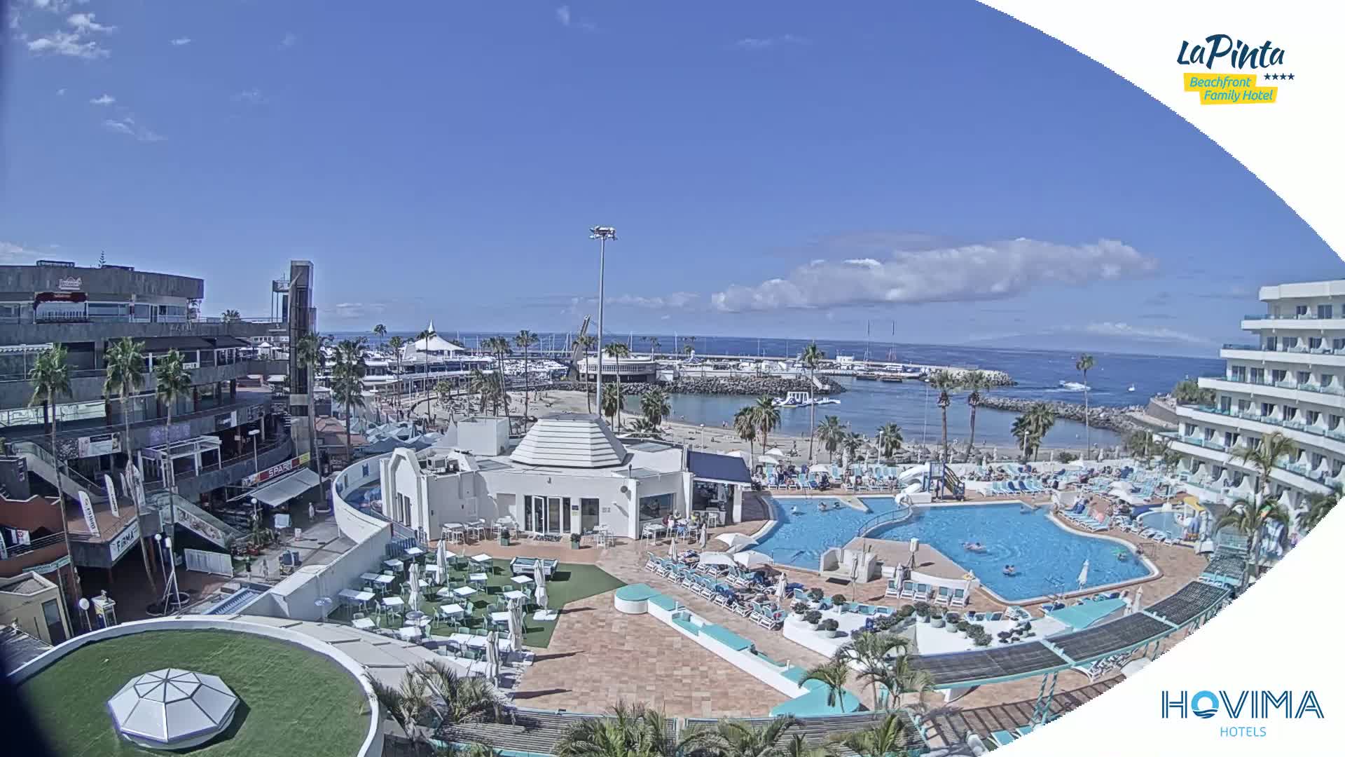 A resort pool area with a marina and ocean in the background under a partly cloudy sky.
