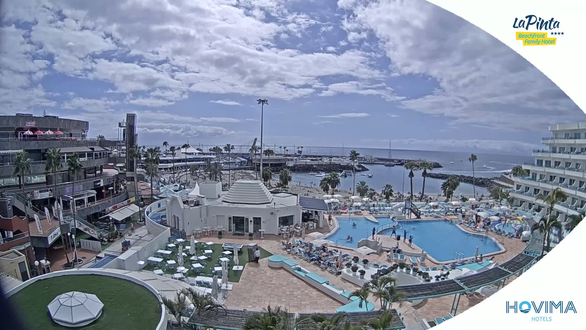 A partly sunny day overlooking a resort with multiple swimming pools, a marina, and shops, near a calm ocean.
