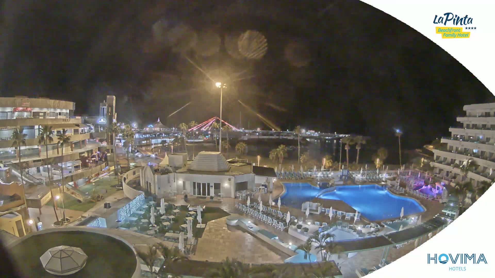 A nighttime view of a resort complex with swimming pools, surrounding buildings, and a beach in the distance under a cloudy sky.