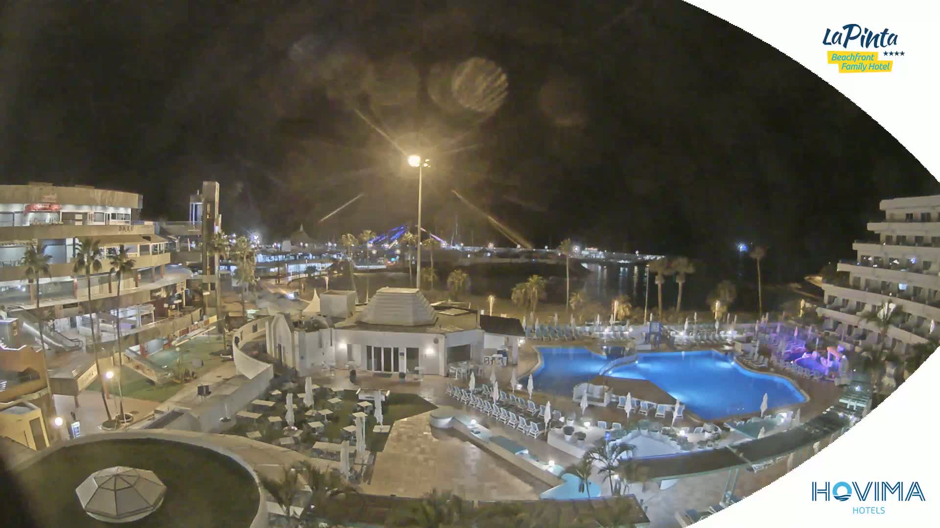 A nighttime view of a resort complex with multiple pools, surrounding buildings, and a waterfront in the distance under a partly cloudy sky.