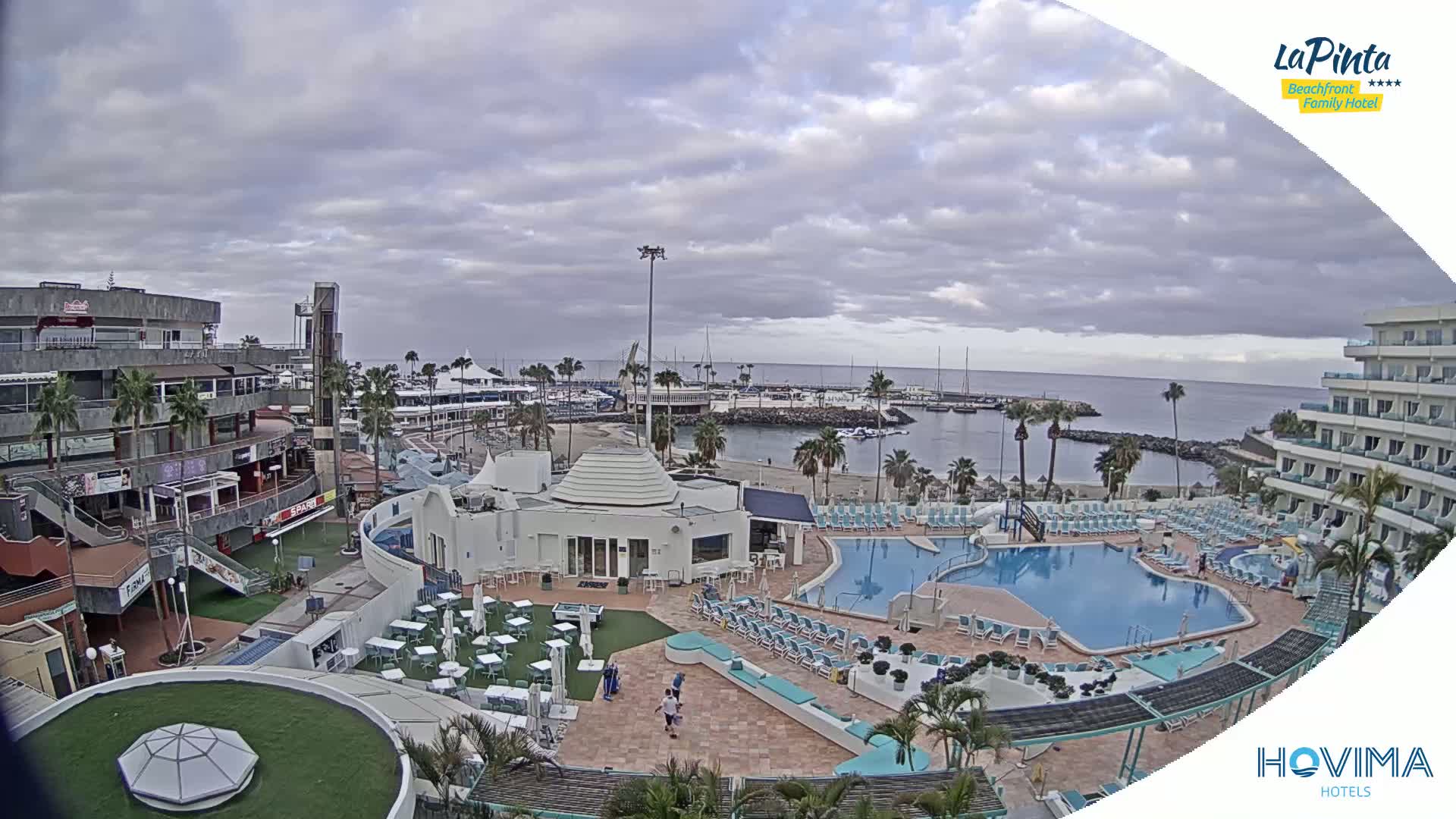An overcast day reveals a resort area with a swimming pool, a small beach, a marina, and various buildings, including shops and hotels.