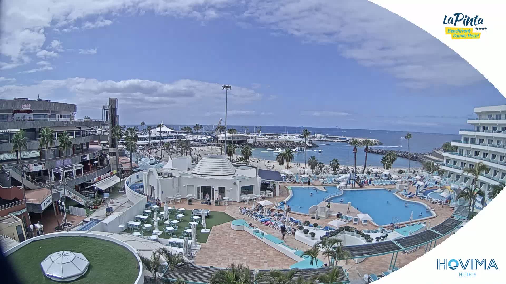 A resort area with a swimming pool and beach, situated next to a marina and ocean, under a partly cloudy sky.