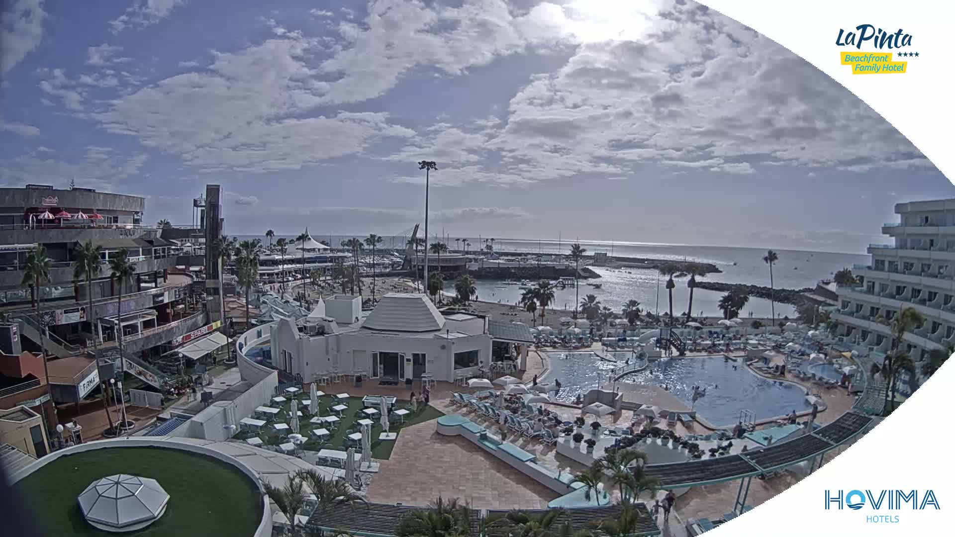 A resort area with a swimming pool, restaurants, and shops overlooks a calm ocean under a partly cloudy sky.