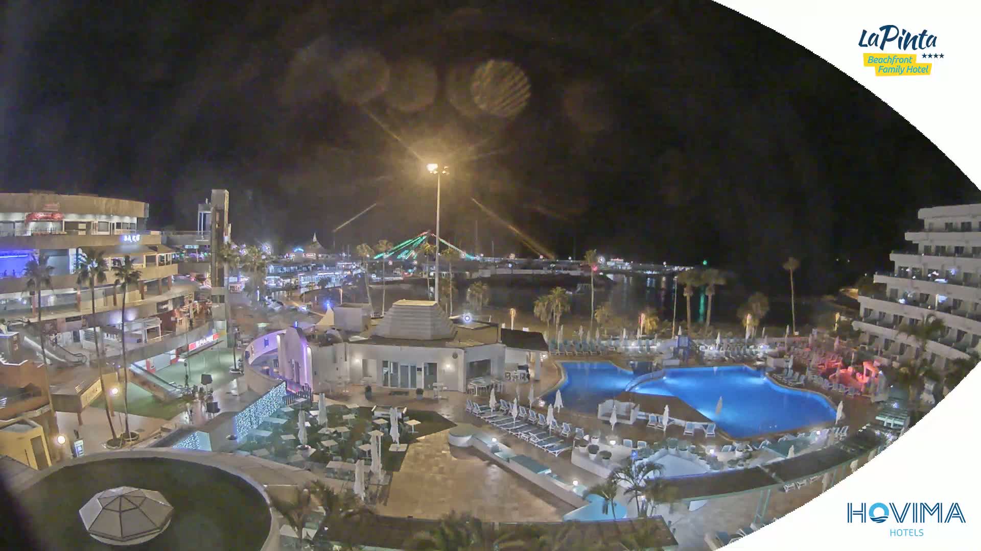 A nighttime aerial view of a resort complex with swimming pools, palm trees, and a brightly lit boardwalk along a calm body of water under a dark, clear sky.