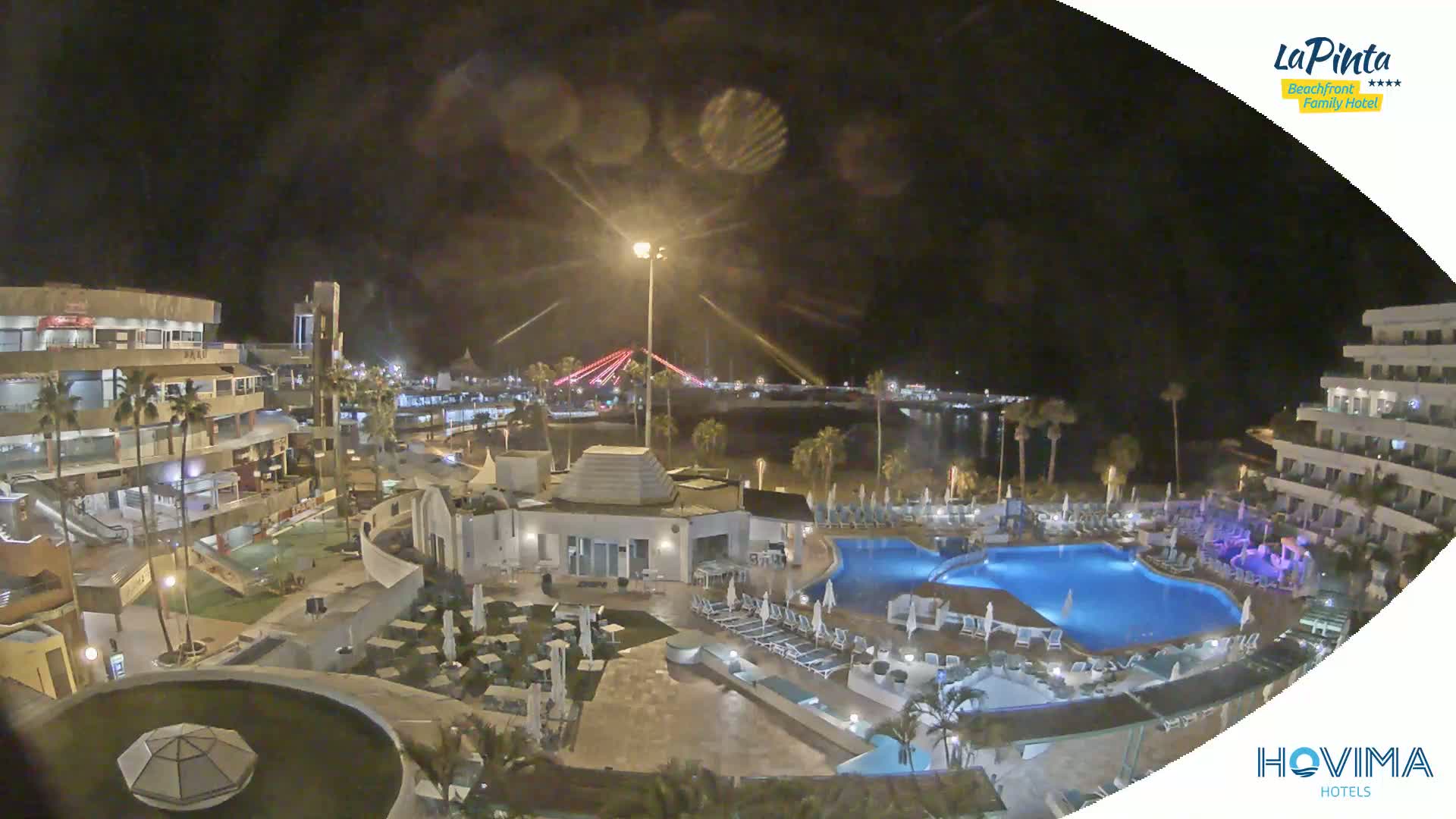 A nighttime aerial view of a resort complex with swimming pools and surrounding buildings, under a slightly overcast sky.