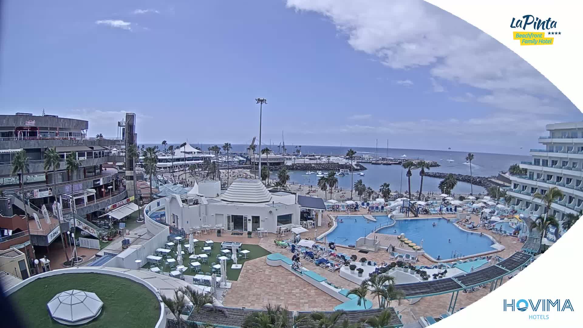 A sunny day overlooking a resort area with multiple pools, a marina, and shops, all situated near a calm ocean.