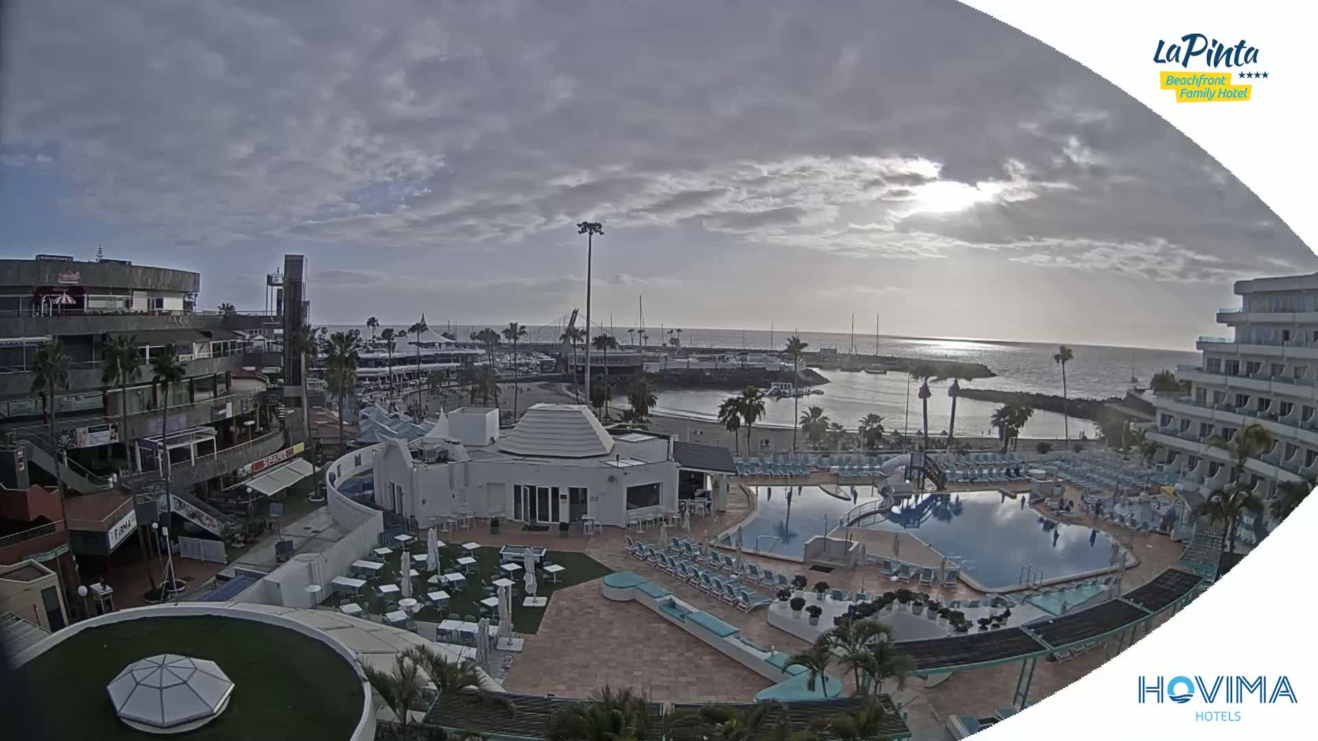 An overcast day reveals a resort area with a marina, pools, and buildings, overlooking a calm ocean.