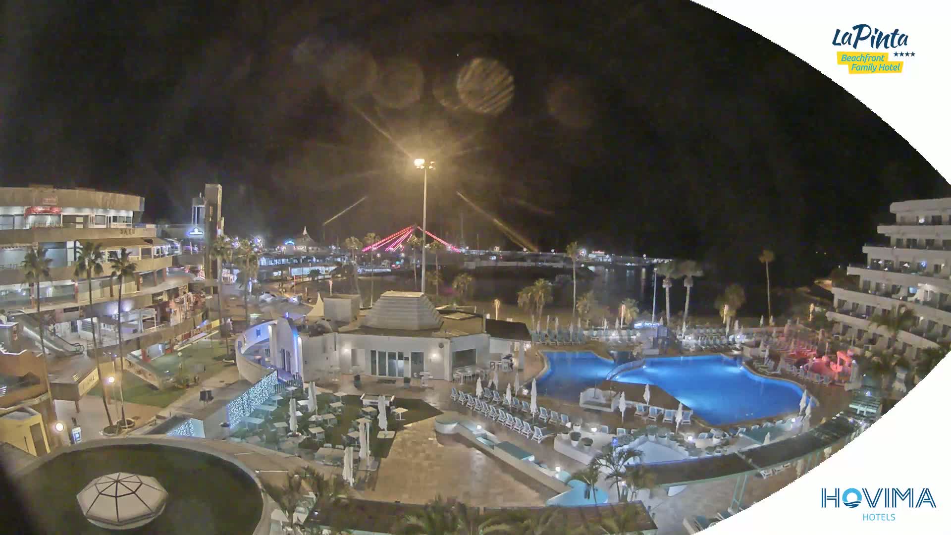 A nighttime view of a resort complex with pools, palm trees, and a brightly lit bridge in the distance under a cloudy sky.