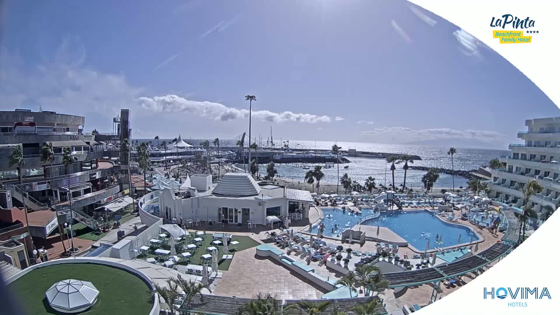 A resort pool area with a beach and ocean view under mostly sunny skies with a few scattered clouds.