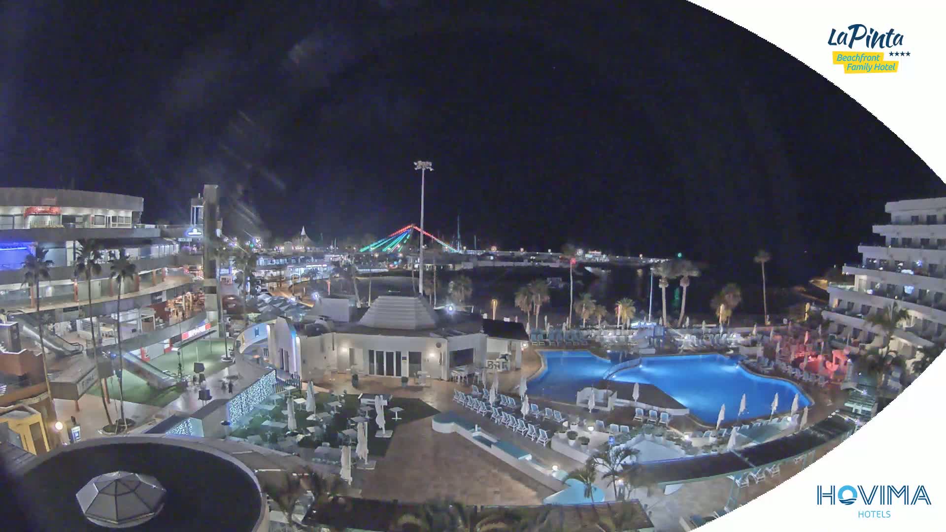 A nighttime aerial view of a resort complex with a swimming pool, illuminated buildings, and a bridge in the distance under a clear, dark sky.