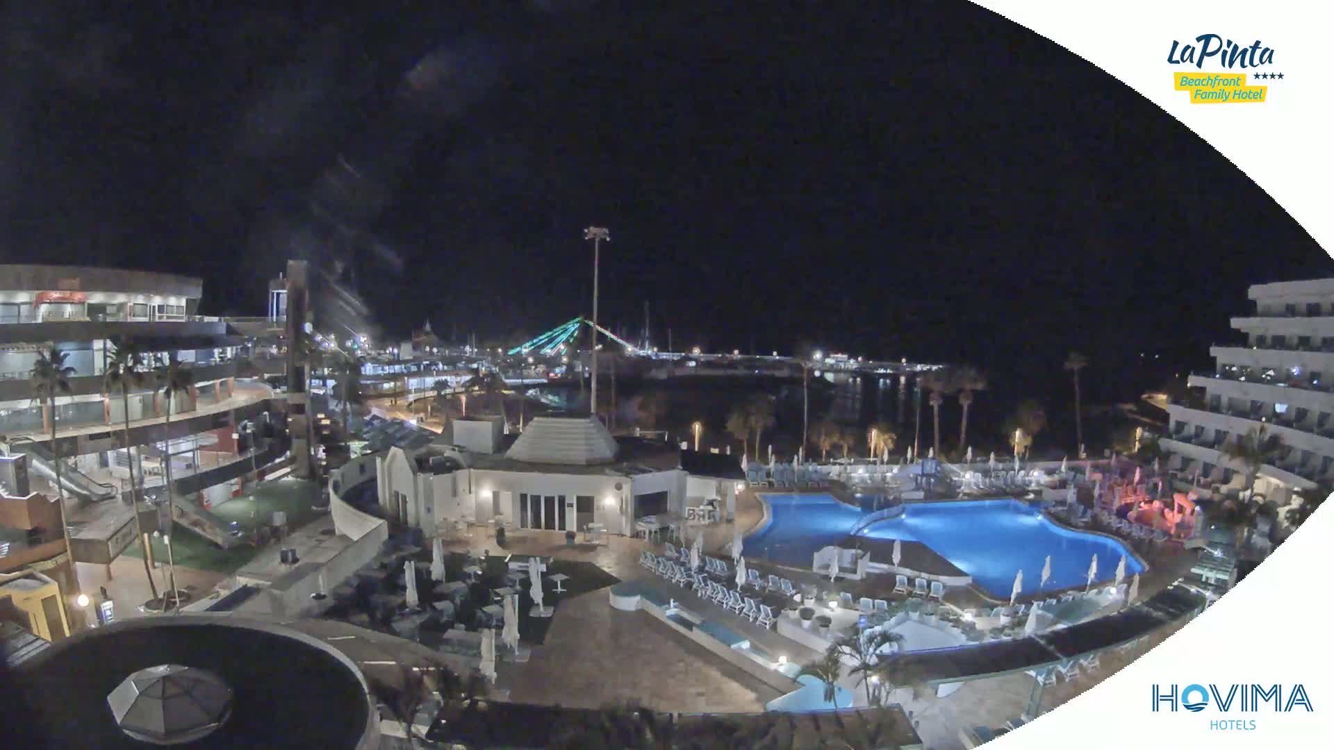 A nighttime view of a resort complex with a swimming pool, illuminated buildings, and a marina with a bridge in the background under a clear, dark sky.