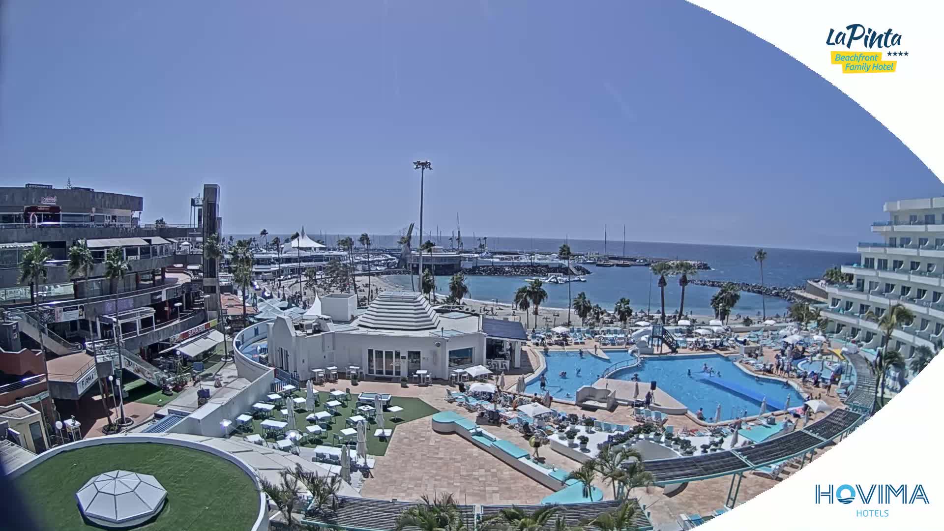 A sunny, beachfront resort with multiple pools, surrounding buildings, and a marina is visible from an elevated viewpoint.