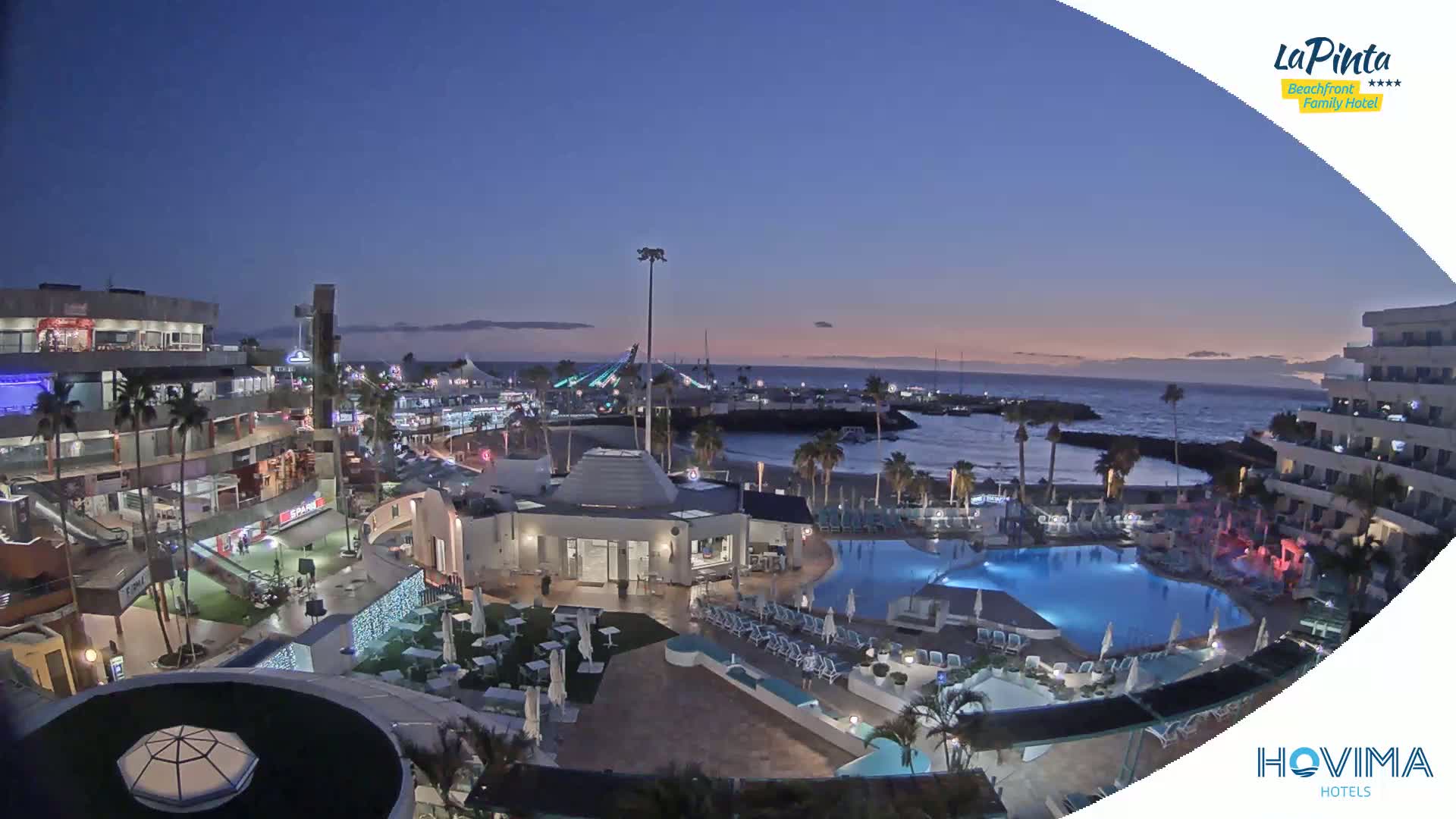 An illuminated resort complex with pools overlooks a calm ocean at twilight.