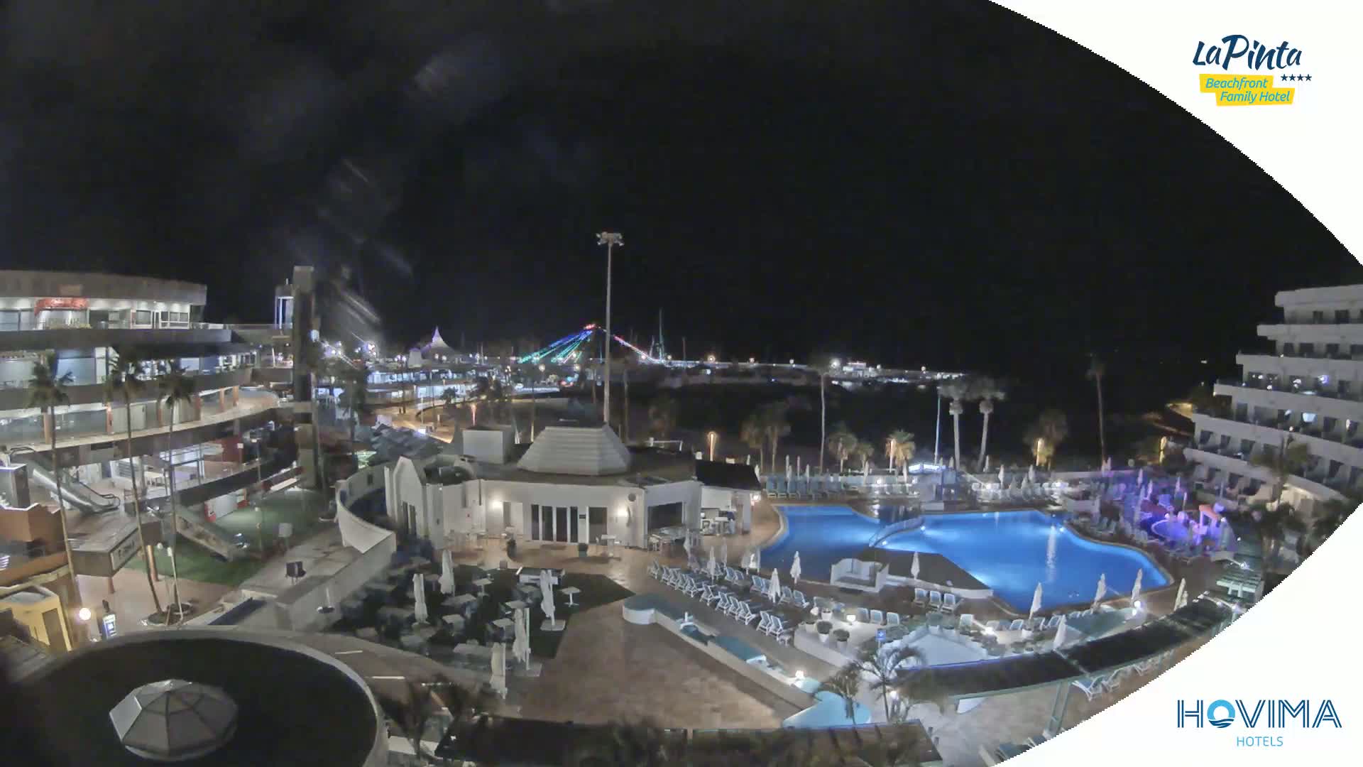 A nighttime aerial view of a resort complex with pools and palm trees, under a clear, dark sky, showing a brightly lit waterfront in the distance.