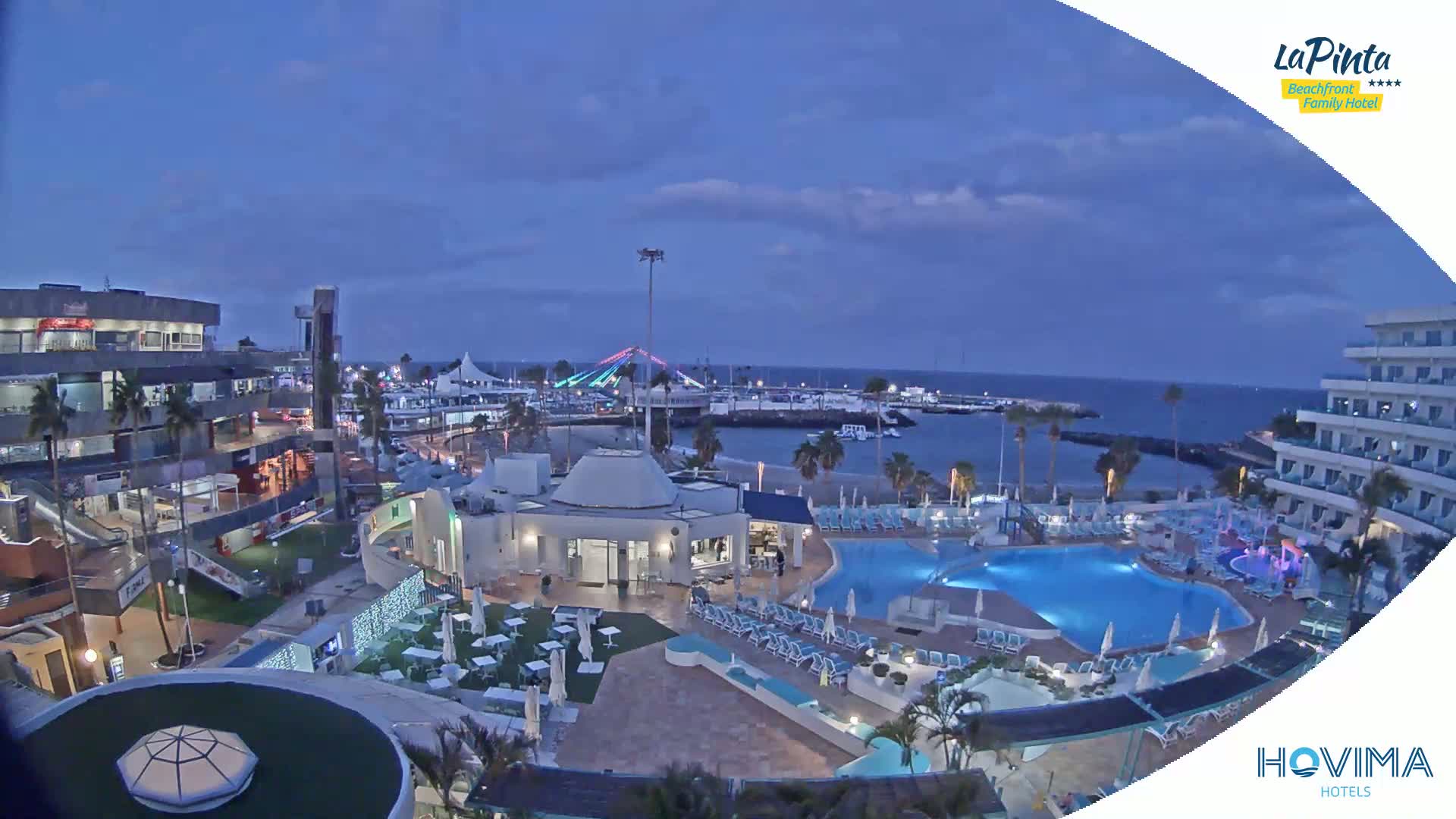 A nighttime aerial view of a resort with multiple pools and a marina, under a twilight sky.