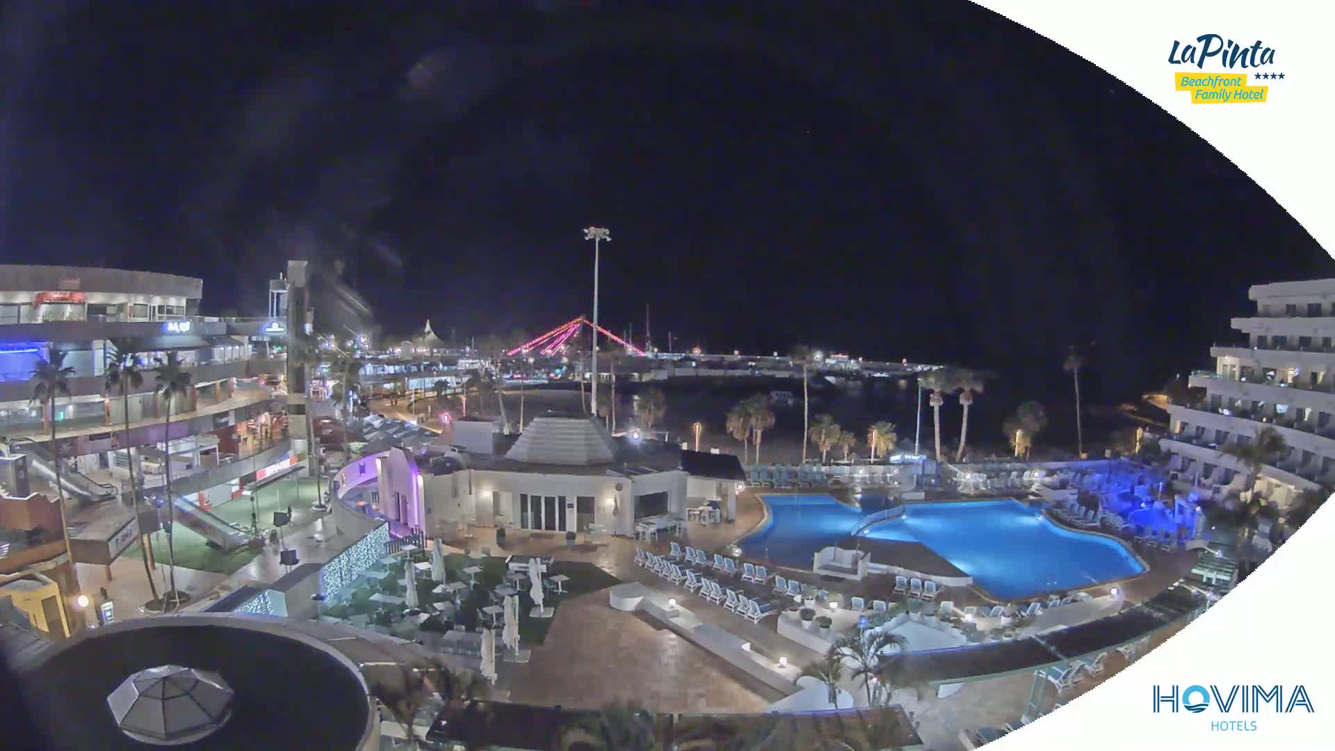 A nighttime aerial view of a resort complex with swimming pools, situated near a waterfront with palm trees and a bridge illuminated with pink lights.