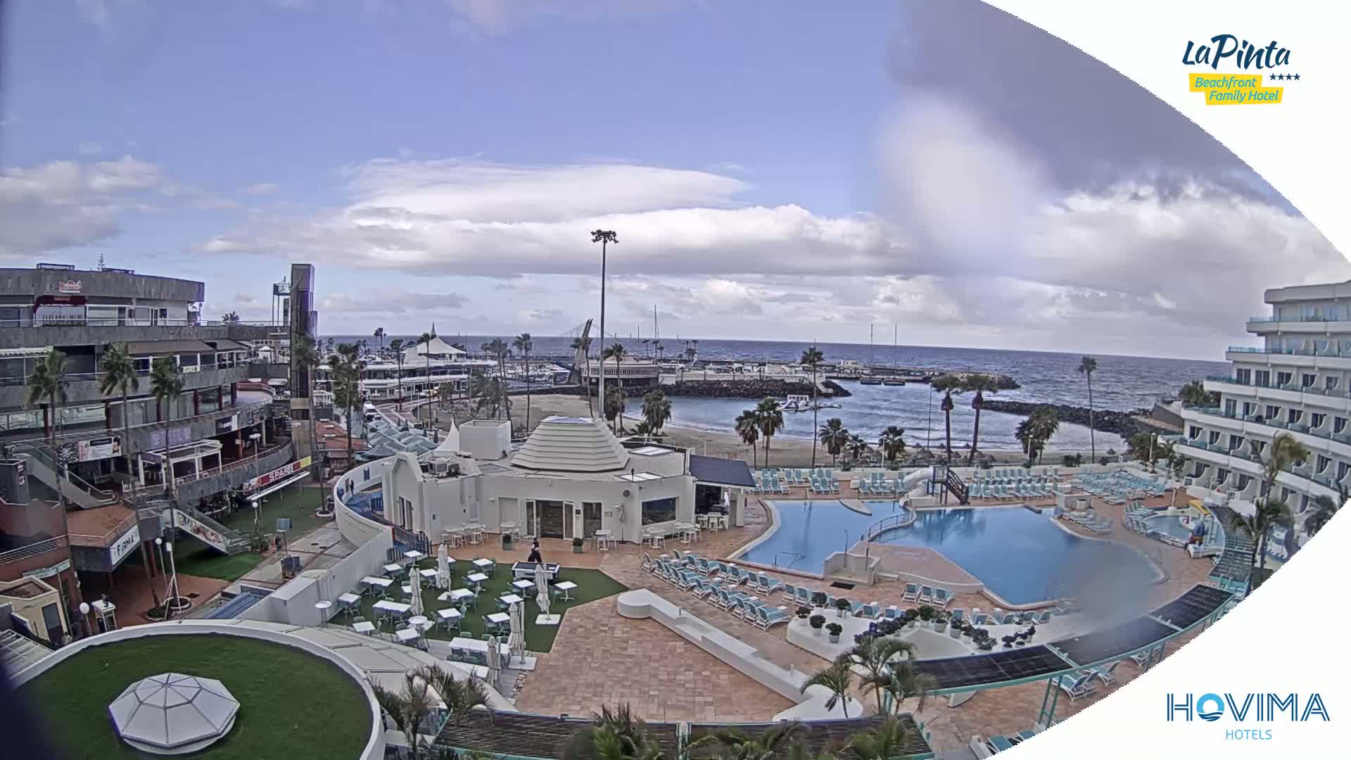 An aerial view shows a resort pool area, adjacent buildings, and a harbor with calm ocean under a partly cloudy sky.