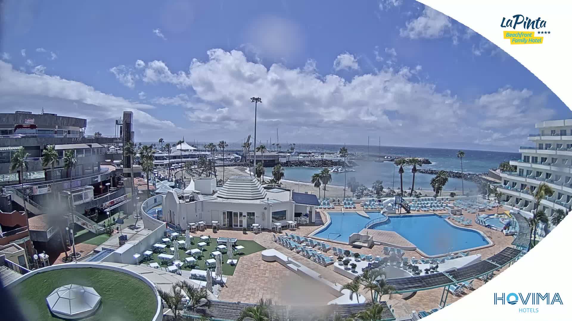 A resort with multiple pools overlooks a calm, partly cloudy ocean harbor with boats and a sandy beach.